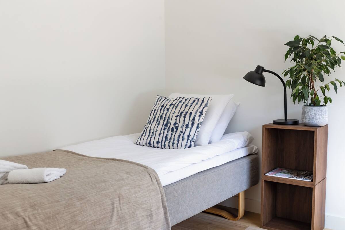 A neatly made bed with a grey blanket and a patterned pillow, a bedside table with a lamp and a potted plant, and a wooden floor.