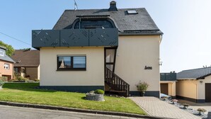 Una casa de dues plantes amb disseny modern, amb un gran finestral a la planta superior i un petit porxo davanter.