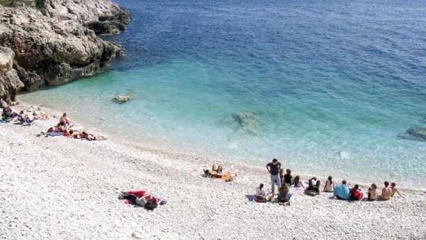 Ein Strand mit klarem türkisblauem Wasser, felsigen Klippen und sonnenbadenden Personen.