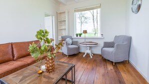 En stue med en brun lædersofa, et glasbord, to grå lænestole, et rundt træbord og et vindue med udsigt til træer.