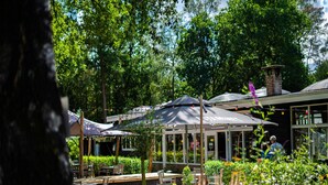 An outdoor seating area with tables and umbrellas, surrounded by lush greenery and trees. - Nature-Connected Modern Stay (Hoenderloo)