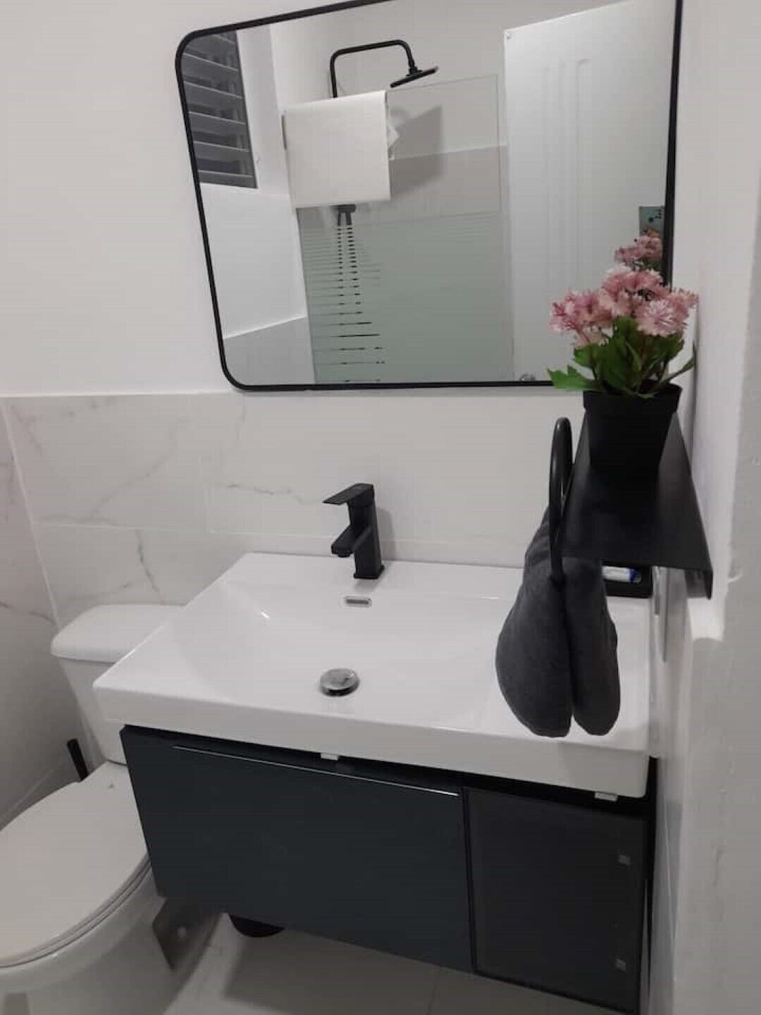 Un baño con un lavamanos blanco, un grifo negro y un espejo encima. Hay una toalla negra colgada en la pared y un florero con flores sobre el mostrador del lavamanos.