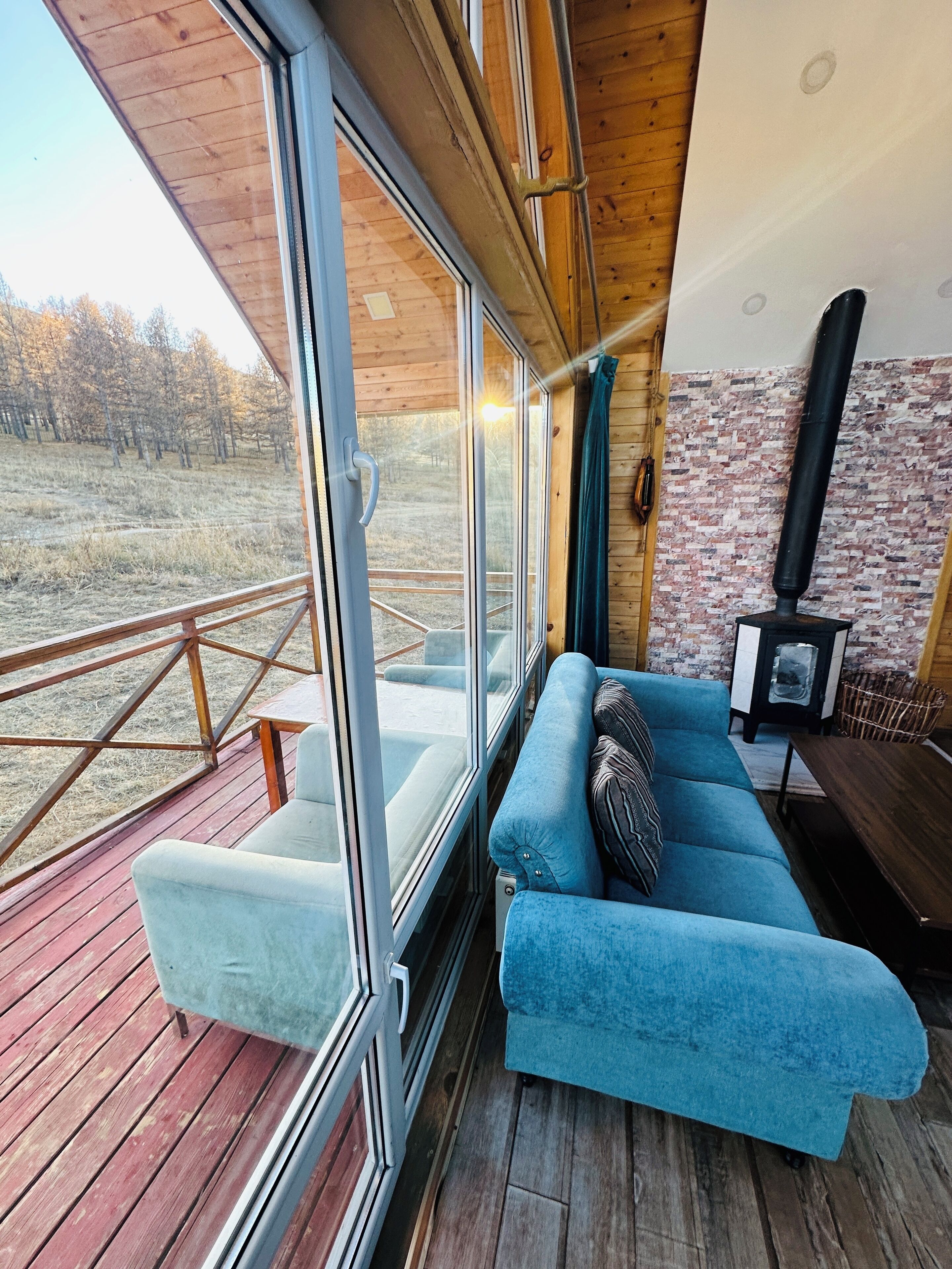 Una habitación acogedora con un sofá azul, una terraza de madera, una chimenea y un ventanal grande con vistas a un campo.