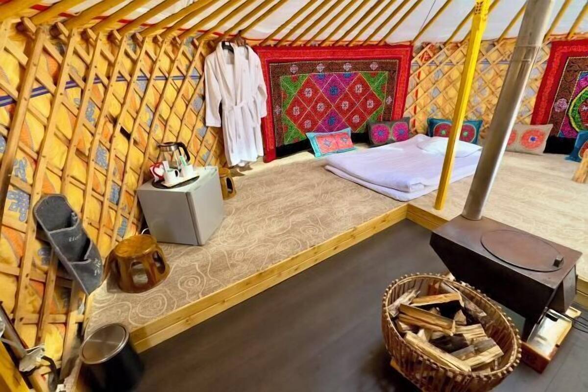 Una habitación con una cama, una mesita pequeña, un taburete, un calentador y un tapiz decorativo.