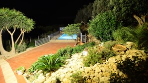 A nighttime view of a landscaped area with a pool, surrounded by various plants and a rocky terrain. - Villa with pool (Trappeto)