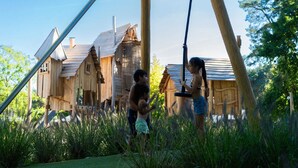 A rustic wooden house with a sloped roof and a chimney, surrounded by tall grass and trees. - Chalet by Pond in Holiday Park (Veldhoven)