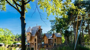 A wooden cabin with a staircase and a slide, surrounded by trees and a sandy area. - Chalet by Pond in Holiday Park (Veldhoven)