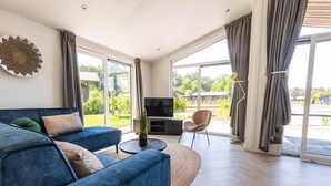 A modern living room with a blue sofa, a flat-screen TV, and a large window with curtains. - Chalet by Pond in Holiday Park (Veldhoven)
