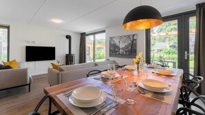 Una sala moderna con un sofá, una mesa de comedor con platos y vasos, un televisor de pantalla plana montado en la pared y grandes ventanas con vistas al exterior.
