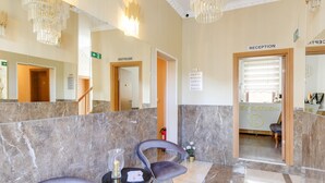 A reception area with marble flooring, two grey chairs, a small table with a glass, and a reception desk. - Apartment in Oberhausen near Lake & River (Oberhausen)