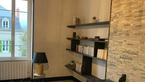 Una sala de estar acogedora con chimenea, estanterías de madera con libros y adornos, una lámpara de pie y vistas a una casa a través de un amplio ventanal.