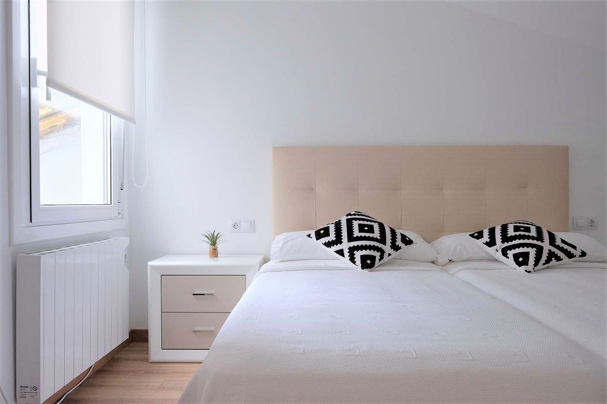 A bedroom with a bed, two pillows, a nightstand, a small plant, and a window with blinds.