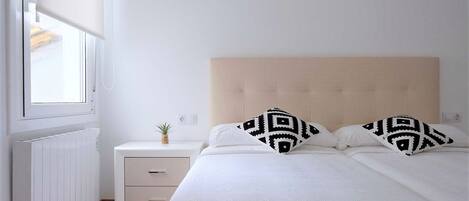 Ein Schlafzimmer mit einem Bett, zwei Kissen, einem Nachttisch, einer kleinen Pflanze und einem Fenster mit Jalousien.