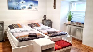 Quarto com duas camas, um radiador, uma janela com persianas, uma mesinha de cabeceira e um quadro pendurado na parede.