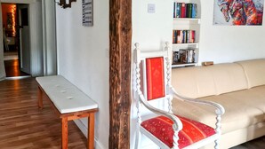 Uma sala de estar com um banco branco, uma poltrona vermelha estofada, um sofá bege e uma estante repleta de livros.