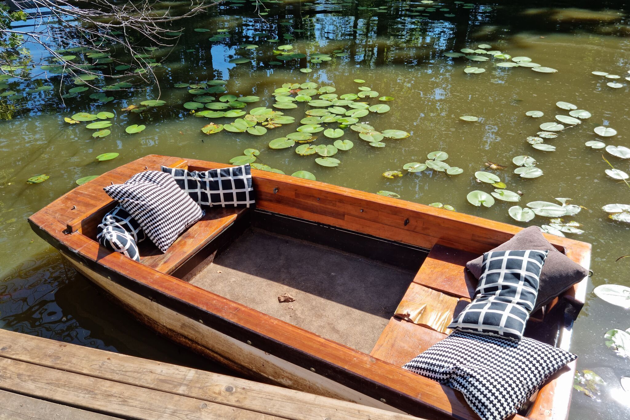 Een houten boot met gestoffeerde zitplaatsen en een vlak oppervlak, afgemeerd aan een houten steiger met waterlelies op het water.