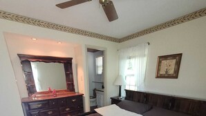Ein Schlafzimmer mit einem großen Bett, einem Deckenventilator, einem Schminktisch, einem Spiegel und einem gerahmten Bild an der Wand.