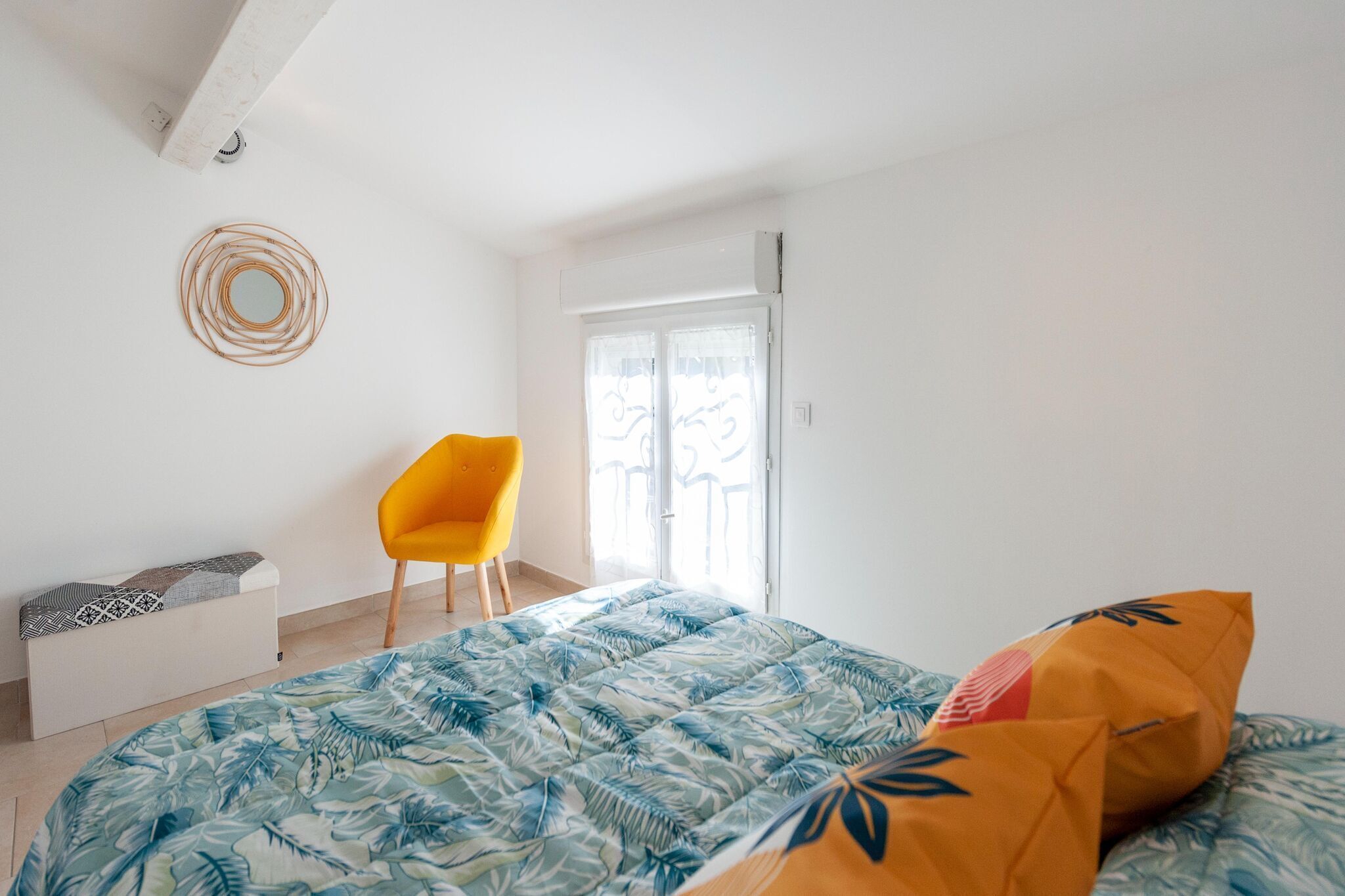 A bedroom with a bed, a yellow chair, a white bench, and a window with a view.