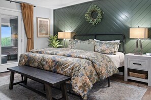 Ein Schlafzimmer mit Bett, Nachttischen, einer Bank, einer Wandlampe, einem gerahmten Bild und Blick durch eine Glastür.