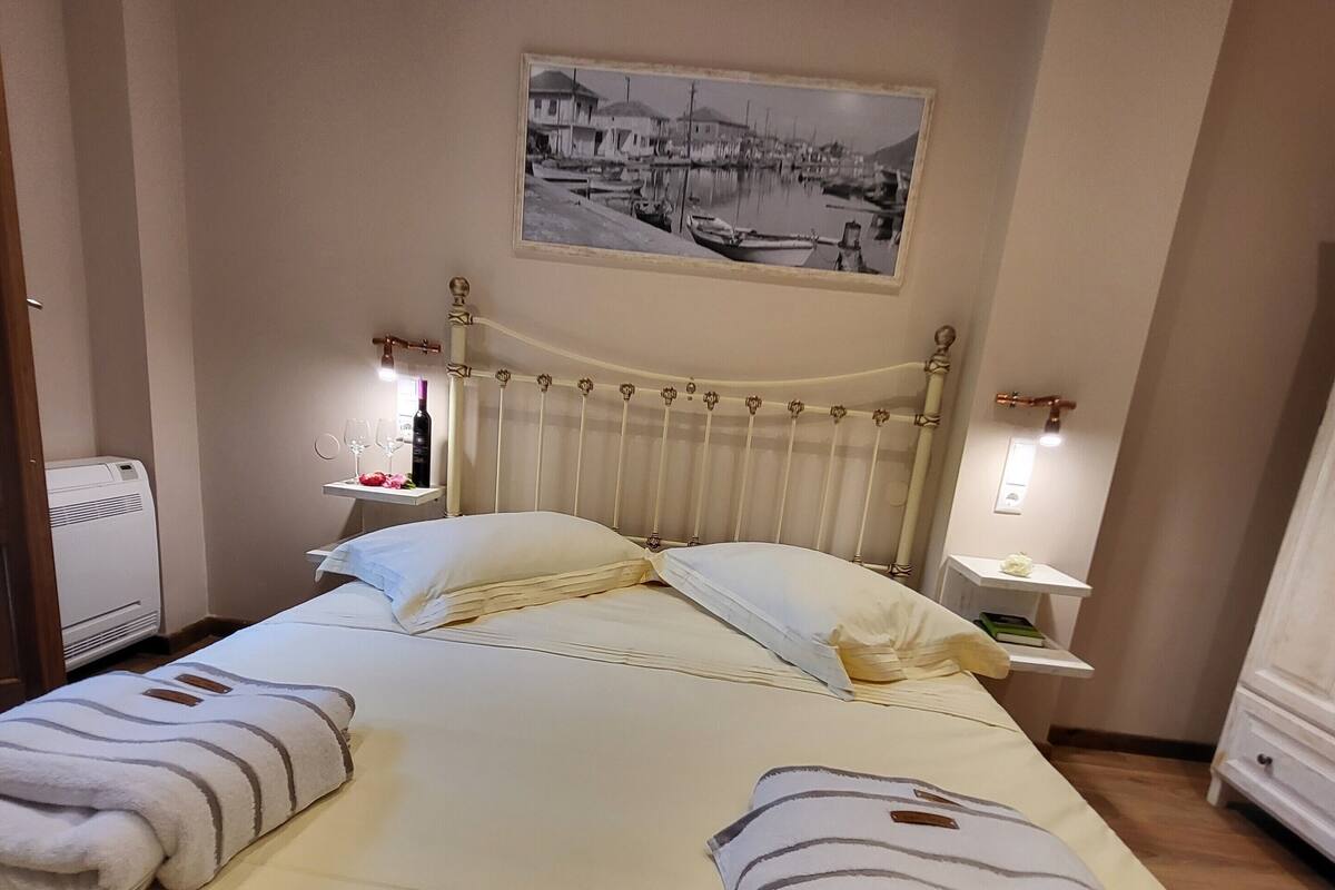 A neatly made bed with pillows and a blanket, a bedside table with a bottle and glasses, and a wall-mounted picture.