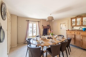 Uma sala de jantar com uma mesa redonda posta para quatro pessoas, rodeada por cadeiras. Há um armário de madeira com portas de vidro à direita, e um lustre sobre a mesa.