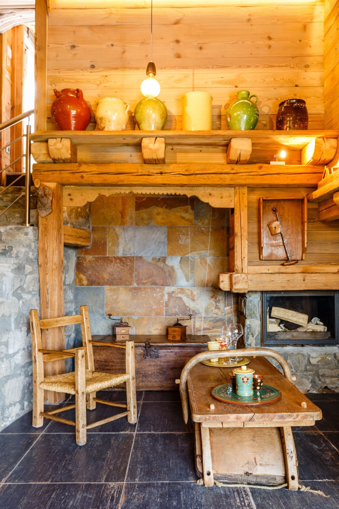 Un interior rústico con chimenea de piedra, estantes de madera con adornos, una mesa de madera con tetera y una silla.