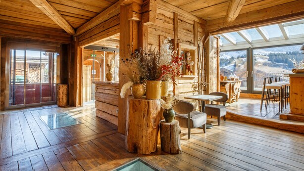 A log cabin interior with wooden floors, a dining area, and a view of a snowy landscape through large windows.