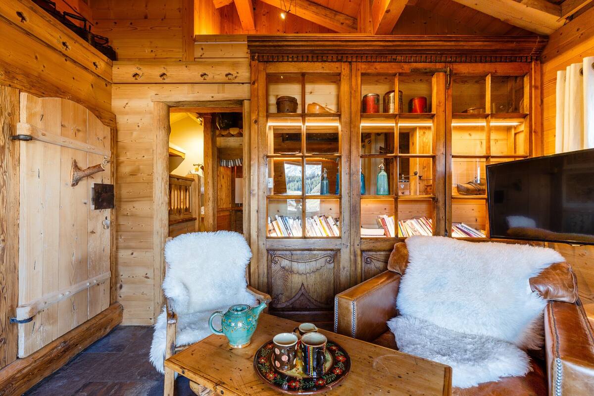 Interior acolhedor de uma cabana de madeira com paredes rústicas, lareira, televisão e estante repleta de livros e objetos decorativos.