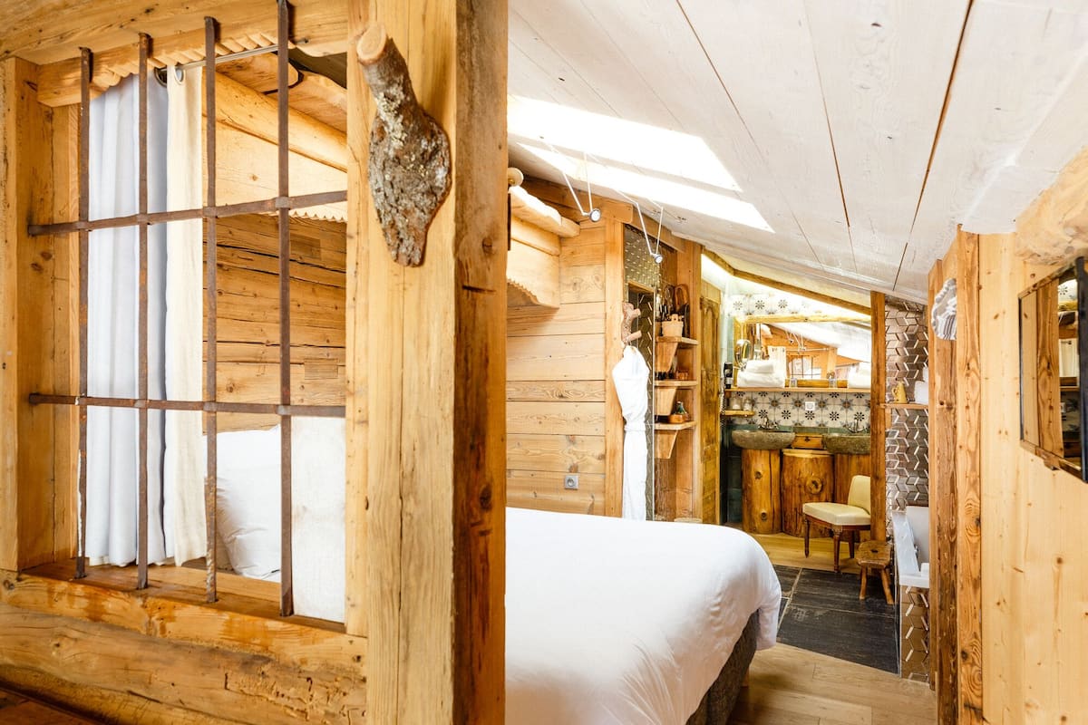 Interior de uma cabana de madeira com uma cama, uma mesa pequena e uma cozinha com pia e prateleiras.
