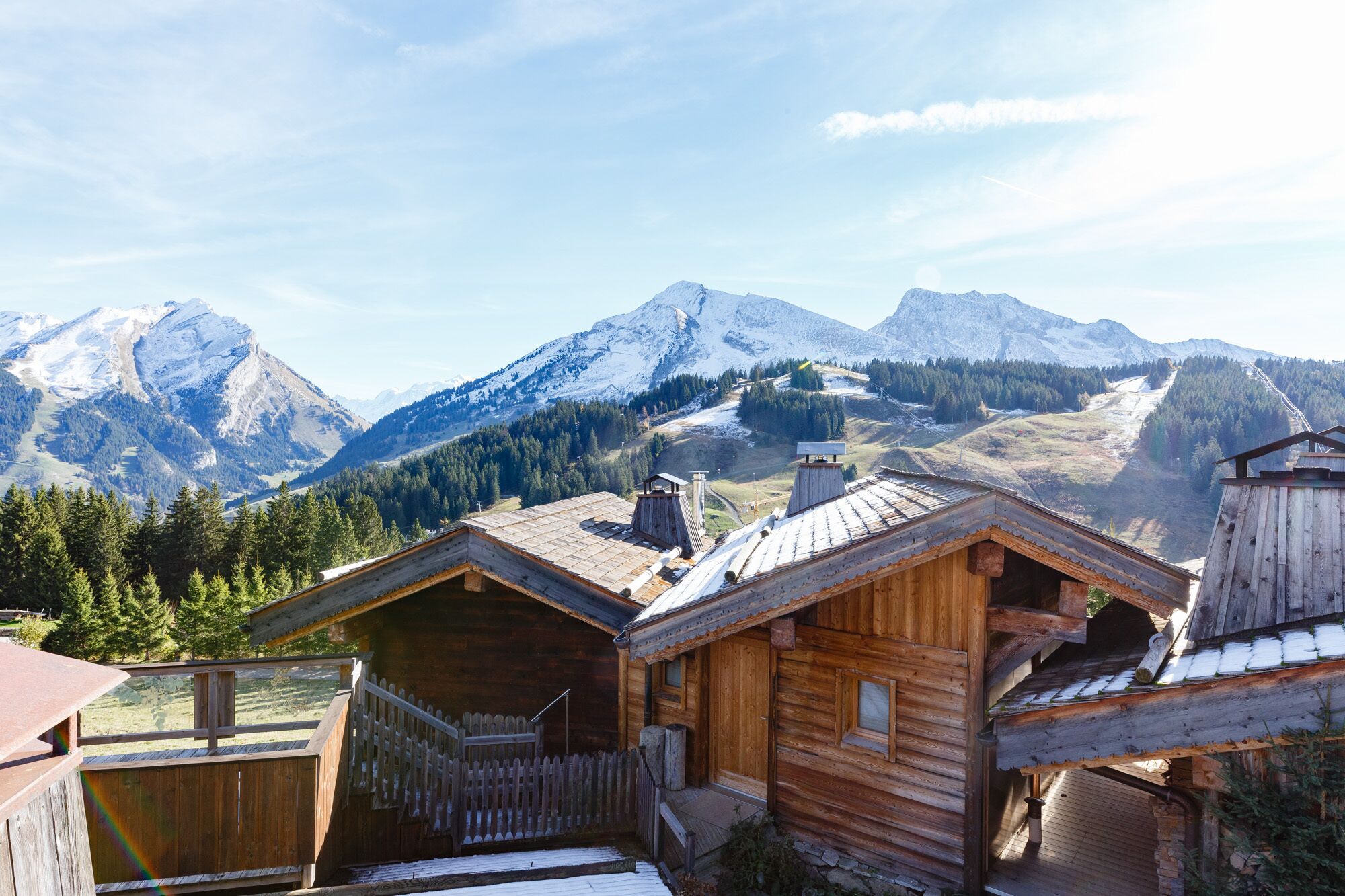 A wooden chalet with a sloped roof, surrounded by a wooden fence, and set against a backdrop of snow-capped mountains and a clear sky.