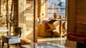 A wooden cabin interior with a wooden table, chairs, a sofa, and a view of a mountain through a large window.