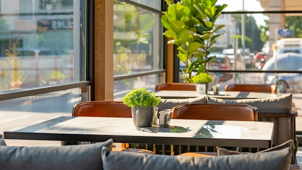 A restaurant with tables and chairs, a potted plant on the table, and a view of the street outside. - Perast City Hotel (Konyaalti)
