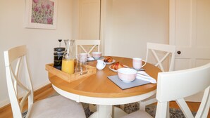 A dining area with a wooden table, white chairs, a coffee maker, a pitcher, glasses, a bowl, a plate with croissants, and a framed floral artwork on the wall. - Holly Leaf Cottage - Drum Castle Estate (Banchory)