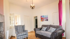 A living room with a grey plaid armchair, a dark brown sofa with grey cushions, a wooden floor, a hanging light fixture, and a painting on the wall. - Holly Leaf Cottage - Drum Castle Estate (Banchory)