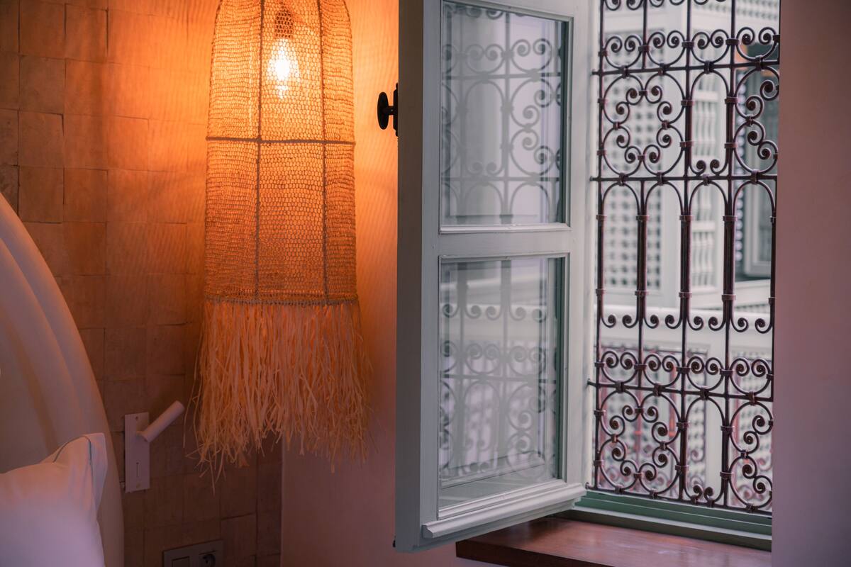 A bathroom with a woven light fixture, a white door with decorative glass panels, and a view of a building through a barred window.