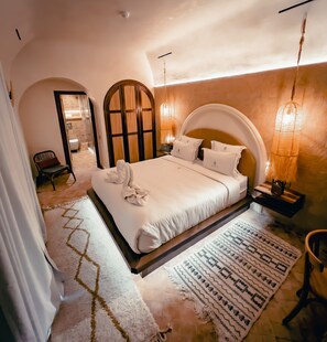 Un dormitorio amplio con una cama grande, una zona de estar con una silla, y un baño visible a través de un arco.