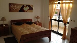 A bedroom with a bed, two bedside tables, a nightstand, a window with curtains, and a wall decoration. - T3 Vivenda Bemposta / Portimão (Portimão)