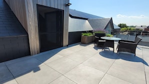 A rooftop patio with a modern design, featuring a large sliding door, a small table with chairs, and potted plants.