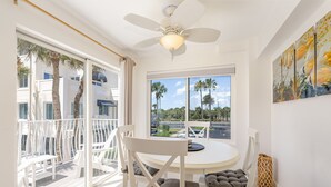 A room with a round table, white chairs, a ceiling fan, a window with a view of palm trees, and a painting on the wall.
