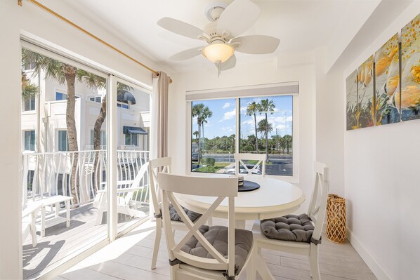 A room with a round table, white chairs, a ceiling fan, a window with a view of palm trees, and a painting on the wall.