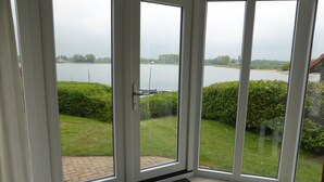 A room with a large sliding glass door leading to a view of a lake and a grassy area with a boat dock. - Chalet Retreat in Green Setting (Lathum)