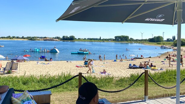 A beach with people swimming and sunbathing, a boat in the water, and a large umbrella providing shade. - Chalet Retreat in Green Setting (Lathum)