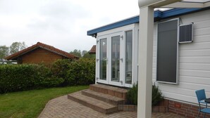 A house with a brick patio, a blue chair, and a white door with glass panels. - Chalet Retreat in Green Setting (Lathum)