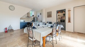 A dining area with a table set for four, a kitchen in the background, and a bookshelf with various items. - Gite in the heart of Saône et Loire (DOMPIERRE LES ORMES)