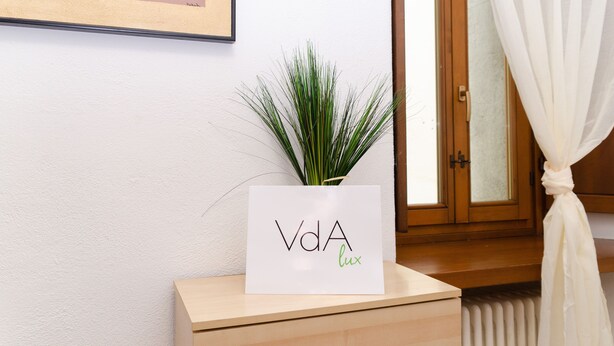 Un panneau blanc avec l’inscription « VdA lux » posé sur une table en bois, une plante verte à côté, et une fenêtre avec des rideaux blancs.