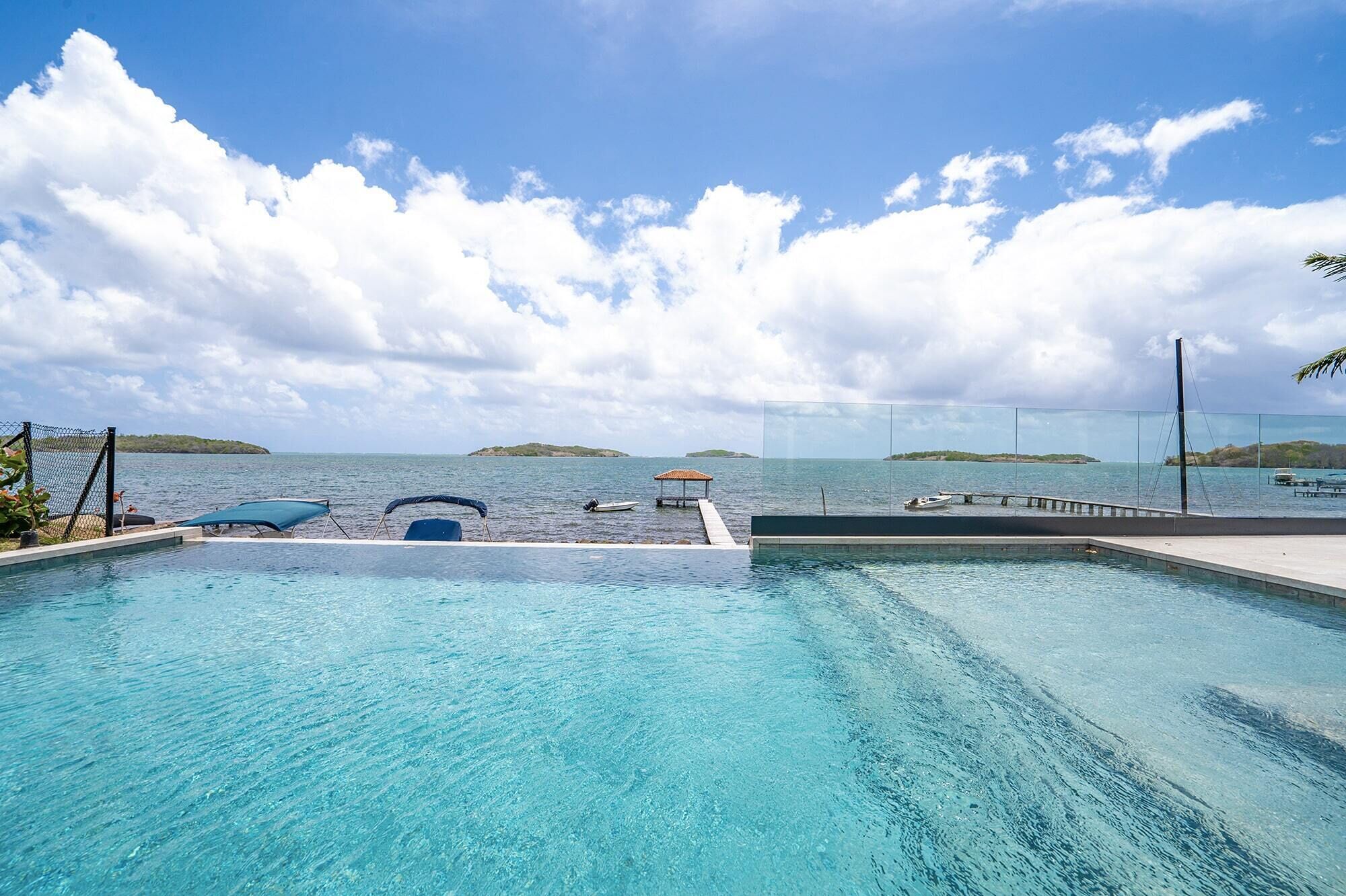 A clear blue pool with a view of the sea and islands in the distance, a covered seating area, and a dock with a boat.