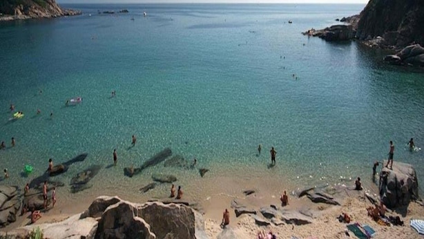 A coastal beach scene with clear water, rocky shore, and people swimming and sunbathing.