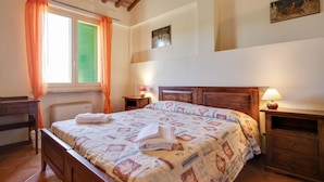 A bedroom with a wooden bed, two bedside tables with lamps, a window with curtains, and a small painting on the wall.