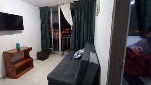 Een woonkamer met een bank, een aan de muur bevestigde televisie, een houten bijzettafeltje en een balkon met gordijnen.