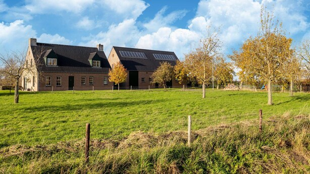 A large brick building with a sloped roof and solar panels, surrounded by a green field and trees. - Eco-friendly apartment with a garden (Sint Kruis)